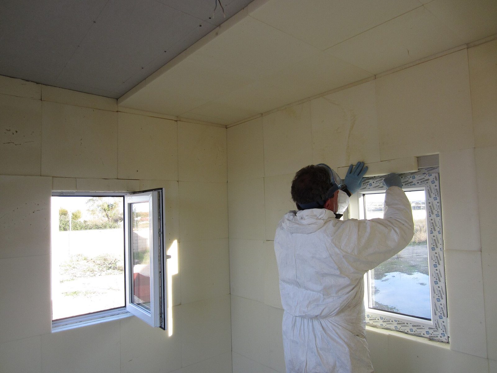 Arbeider in een witte overall werkt de isolatie af rond een nieuw geplaatst kunststof raam in een ruwbouwkamer