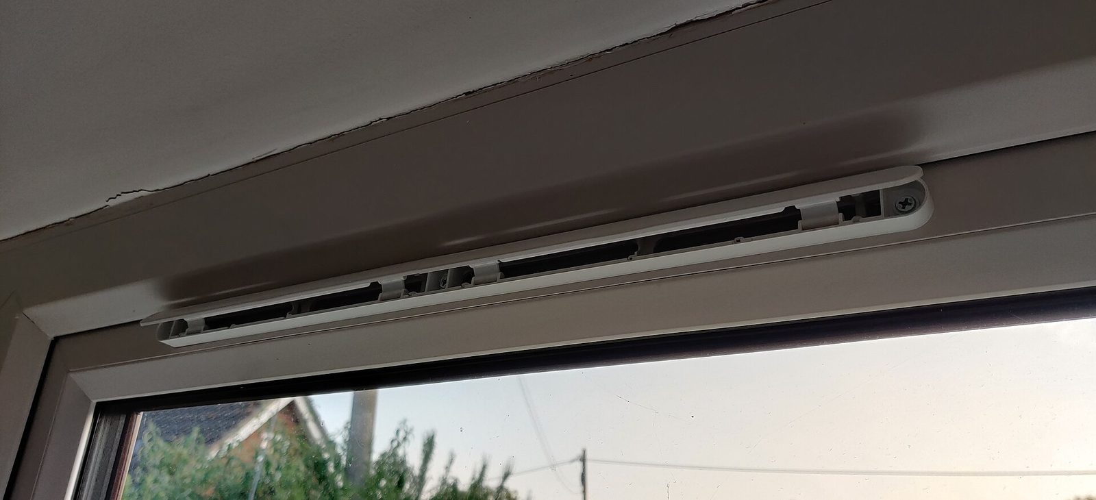 Geopend ventilatierooster bovenin een wit raamkader, met daaronder zicht op een dak en de lucht