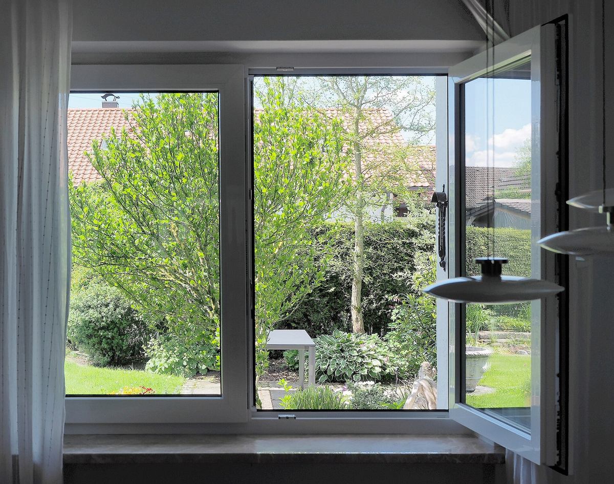 Wit raam met een openstaande vleugel die uitkijkt op een groene tuin met struiken en de daken van de buren