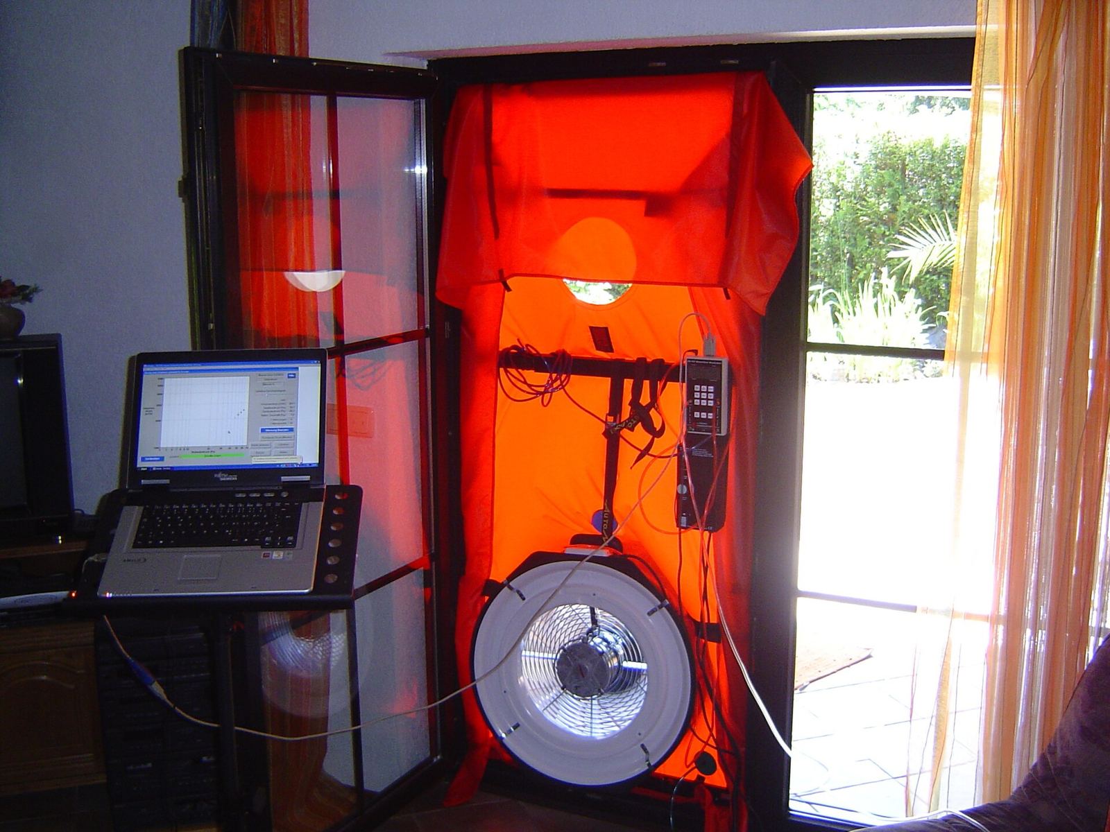 Blowerdoortest in een terrasdeur: oranje doek met ventilator en meettoestel, aangesloten op een laptop